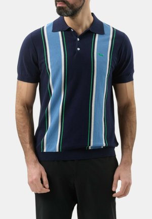 Uomo che indossa una polo blu navy a maniche corte con righe verticali azzurre, bianche e verdi, abbinata a pantaloni neri.