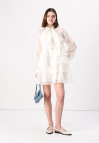 Rochie albă în straturi, cu mâneci lungi din material transparent și o fundă la gât. Pur și simplu cu pantofi bej și o mică poșetă albastră. Textură netedă.