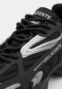 Zapatilla negra con material de malla y goma. Presenta acentos plateados, superposiciones texturizadas y cordones prominentes. Marca Lacoste visible.