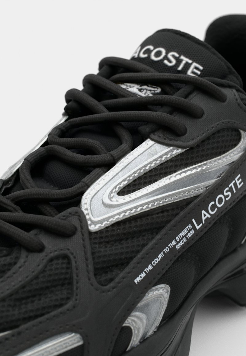 Baskets noires en matériau en mesh et en caoutchouc. Dotées d'accents argentés, de superpositions texturées et de lacets proéminents. Marque Lacoste affichée.