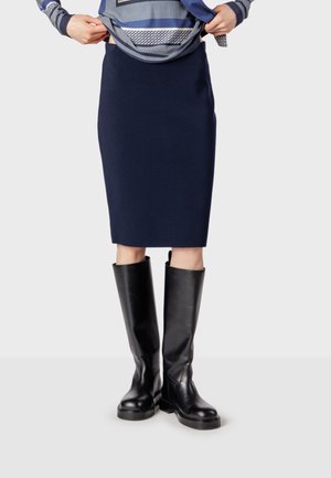 Personne portant une jupe bleu marine mi-genoux, remontant un top bleu à motifs, et portant des bottes en cuir noires montantes jusqu'aux genoux.