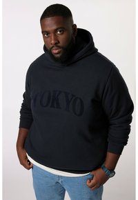 Felpa navy con testo "TOKYO" in blu scuro, tessuto misto cotone, tasca a marsupio, indossata sopra una camicia bianca, abbinata a jeans azzurri.