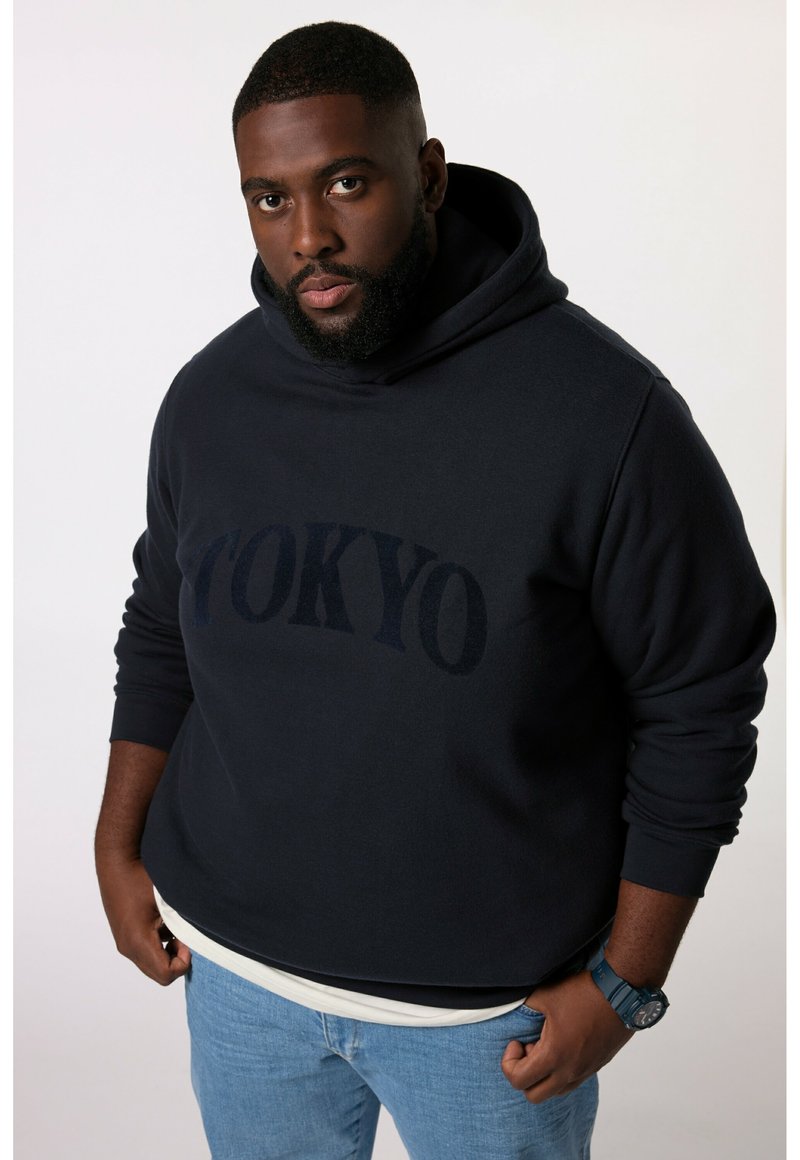 Felpa navy con testo "TOKYO" in blu scuro, tessuto misto cotone, tasca a marsupio, indossata sopra una camicia bianca, abbinata a jeans azzurri.