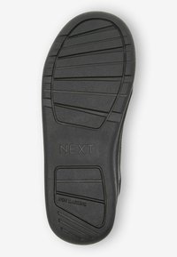 Suola di scarpa in gomma nera con linee di grip testurizzate e il marchio in rilievo "NEXT." Caratteristica non lasciato segni indicata sul lato.