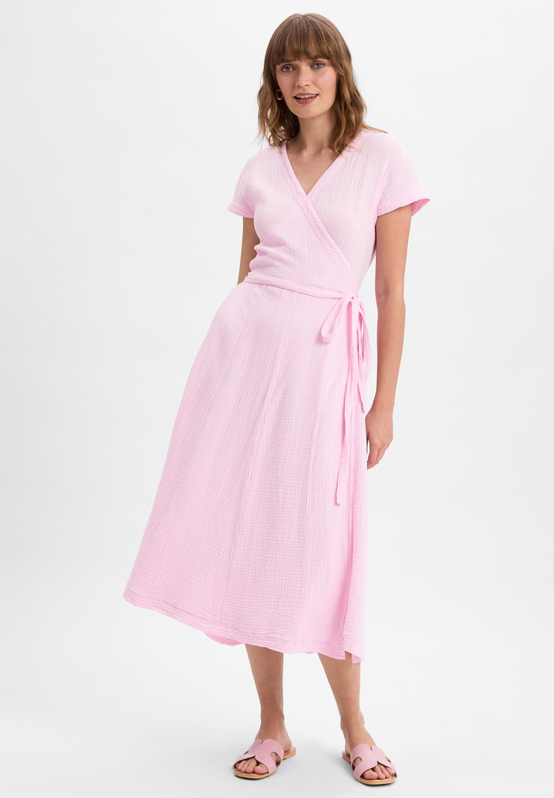 Lichtroze katoenen wikkeljurk met korte mouwen, gestructureerde stof en een strik taille. Gecombineerd met roze slippertjes.