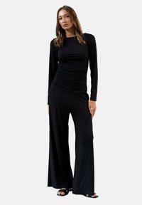Czarny top z długim rękawem o marszczonej fakturze i czarne spodnie z szerokimi nogawkami, oba wykonane z lekkiego materiału. Uzupełnione o czarne sandały na płaskiej podeszwie.