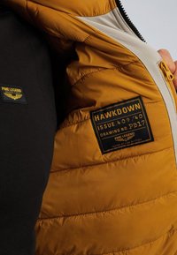 Žlutá péřová bunda s černou podšívkou, se výrazným logem a zipovou kapsou, hladká textura a prošívaný design.
