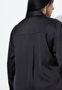 Sort satinskjorte med en glat tekstur, der har en læg bagpå og en klassisk krave. Detaljerne inkluderer en diskret glans og en skræddersyet pasform.