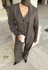 Hnedý oblek na mieru s hlbokým výstrihom do V, tromi gombíkmi a širokými nohavicami. Doplněný zodpovedajúcou šatkou a čiernymi špicatými podpätkami.