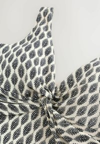 Valge kangas musta lehekujundusega, kokku kootud ja sõlmitud, näidates õlalinte ja tekstuurset mustrit lähivõttes.