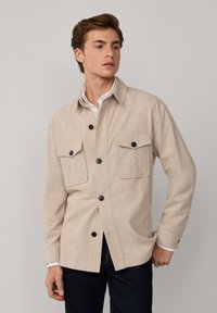 Cazadora beige de botones fabricada en tejido suave, con dos bolsillos en el pecho, botones negros y cuello. Llevada sobre una camisa blanca.