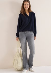 Marineblå bluse med lange ærmer og V-hals, parret med lysegrå jeans. Modellen holder en cremefarvet crossover-taske og har hvide sneakers på.
