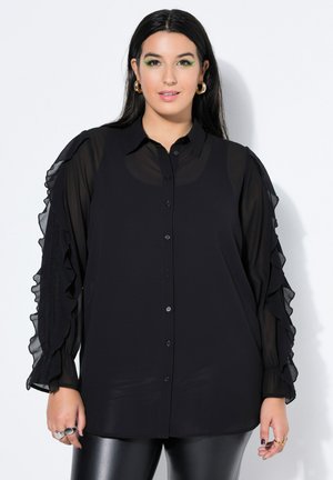 Frau mit langen dunklen Haaren, die eine schwarze transparente Bluse mit Rüschenärmeln und glänzende schwarze Leggings trägt, steht vor einem hellen Hintergrund.