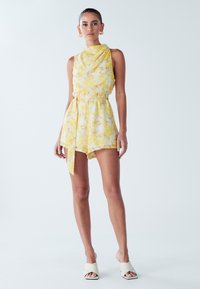 Gele bloemenromper met een hoge hals, een taille die is opgesnoerd met een strik, en ontspannen short. Gladde stof met een lichte textuur.