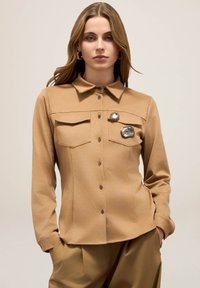 Chemise à manches longues en tissu beige doux avec un col, deux poches poitrine et des broches décoratives sur le côté gauche.