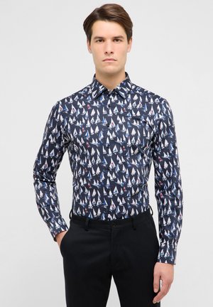 Ung mand iført en marineblå skjorte med små sejlbådsmønstre, matchende med sorte bukser, stående mod en ensartet lys baggrund.