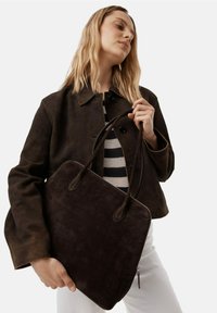 Torba typu tote z ciemnobrązowego zamszu z podwójnymi uchwytami, zamknięciem na zamek błyskawiczny i gładką fakturą, zestawiona z ciemną kurtką i paskowaną bluzką.