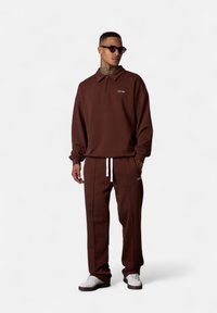 Pull brown avec un col zippé, arborant un logo sur la poitrine. Associé à un pantalon marron assorti avec des cordons blancs, complété par des baskets blanches.