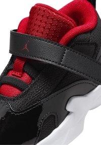 Czarno-czerwony but sportowy z teksturowaną siateczką, błyszczącymi akcentami z lakierowanej skóry i paskiem na rzep nad sznurówkami. Biała podeszwa.