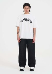 Hvid oversized t-shirt med sort "Ed Hardy" skrift, parret med sorte brede bukser og hvide sneakers. Simpelt design, afslappet pasform.