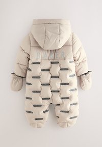 Beige talvivaltio, jossa on huppu, useita mustia ja valkoisia brändättyjä läpysköitä, tikattu pinta sekä elastiset hihansuut ja lapaset.
