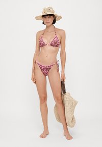 Zestaw bikini z różowym kwiatowym wzorem na brązowym tle; trójkątny biustonosz i majtki z wiązaniem po bokach. Modelka niesie beżową plecioną torbę i nosi słomkowy kapelusz.