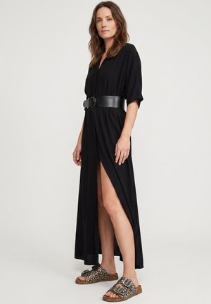 Kobieta stoi w czarnej sukience z paskiem, z rozcięciem z boku i wzorzystych, otwartych sandałach na jednolitym tle.