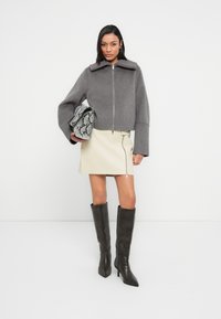 Γκρι cropped σακάκι από μίγμα μαλλιού με φαρδιά μανίκια και ψηλό γιακά, συνδυασμένο με μπεζ μίνι φούστα από δέρμα με πλαϊνό φερμουάρ και καφέ μπότες πάνω από το γόνατο.