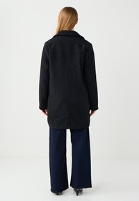 Schwarzer Fleece-Mantel mit tailliertem Kragen, langen Ärmeln und geradem Schnitt, kombiniert mit weit geschnittenen, dunkelblauen Jeans. Glatte Textur insgesamt.