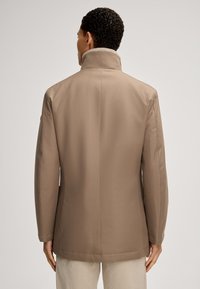 Beige Mantel mit hohem Kragen, glattem Stoff und taillierter Passform. Auffälliges Detail ist die dezent strukturierte Oberfläche und die sauberen Nähte.