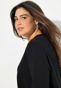 Musta pitkähihaisesti paita, jossa on pyöreä pääntie; malli, jolla on päässään hopeiset renkaat korvakorut ja hiukset valuivat olkapäiden yli, taustalla valkoinen tausta.