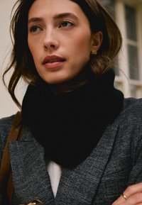 Crna pletena šal nošena uz sivu teksturiranu blejzeru s klasičnim ovratnikom i smeđom trakom, ističući glatki materijal i elegantan dizajn.