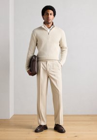 Beige strikkegenser med halvlås, kremfarget skjorte, lysebeige bukser, brun lærmappe og mørkebrune sko, stående mot en enkel vegg.