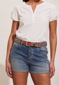 Weißes kurzärmliges Blusenshirt mit Knopfleiste und gerafften Schultern, kombiniert mit blauen Jeansshorts. Brauner, mit Nieten verzierter Gürtel mit einer Metallschließe.