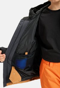 Wattierte Jacke mit grauem Außenmaterial, schwarzem Innenfutter und orangefarbenen Akzenten. Verfügt über eine Netzinnentasche mit einem blauen Gegenstand darin.