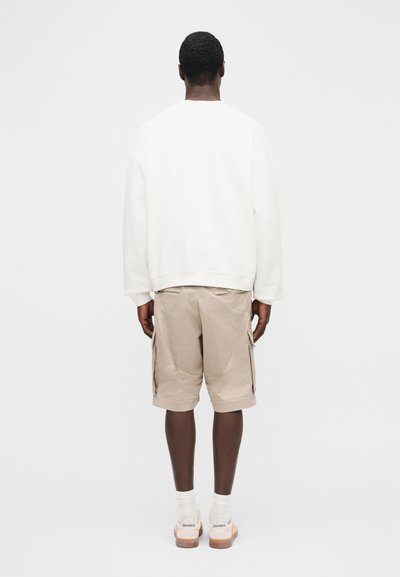 Άνδρας με την πλάτη προς το μέρος του, φοράει λευκό φούτερ, μπεζ σορτς καρό, λευκές κάλτσες και λευκά αθλητικά παπούτσια με λογότυπο της μάρκας στις φτέρνες.