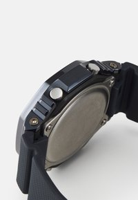 Ceas cu brățară din cauciuc negru, având o suprafață texturată, accente metalice și un capac rotund din argint. Include două butoane și un design detaliat.