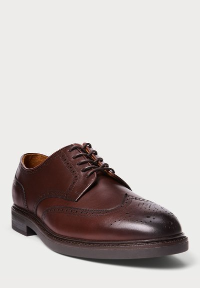 Smešane smeđe kožne cipele s krilima (wingtip brogue) sa tamnosmešnim vezicama, detaljnim perforacijama i složenom petom na bijeloj pozadini.