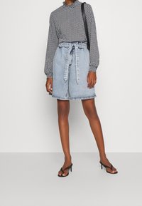 Žena oblečená v dlouhém rukávu topu s černo-bílým houndstooth vzorem, světle modrých džínových šortkách s opaskem, černých sandálech na podpatku a nese černou kabelku přes rameno.