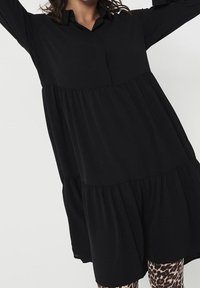 Abito camicia nero a maniche lunghe con colletto abbottonato, strati arricciati e silhouette fluida. Abbinato a leggings con fantasia.