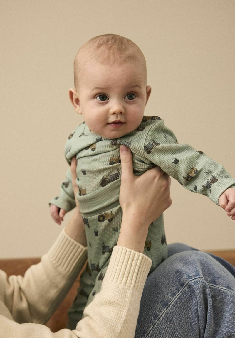 Un vêtement pour bébé à motifs verts représentant des tracteurs, fabriqué en tissu texturé. Tenu par une personne en pull léger, assise sur une surface marron.