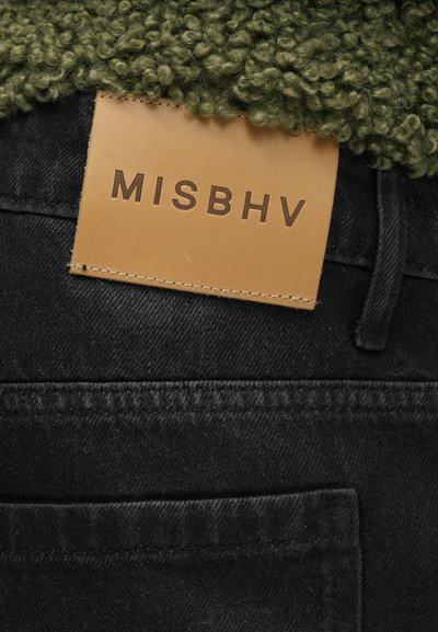 Crne traperice s vidljivom kožnom etiketom utisnutom s natpisom "MISBHV." Debela zelena umjetna krzna tekstura može se vidjeti iznad pojasa.