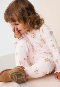 Roosad puuvillased pidžaamad väikeste lillemustritega. Laps istub heledal pinnal, jalas beežid saapad tekstureeritud talladega.