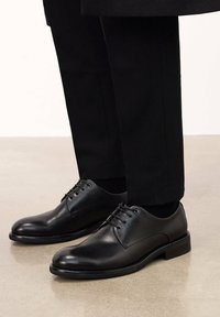 Pantofi de piele negri, cu vârful rotund, cinci găuri pentru șireturi și șireturi plate. Talpa are un finisaj mat neted, care completează perfect pantalonii negri, croiți.