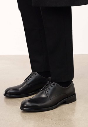 Zapatos de vestir de cuero negro con punta redondeada, cinco ojales y cordones planos. La suela tiene un acabado mate suave, complementando los pantalones negros a medida.