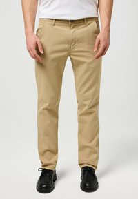 Beige Baumwollhose mit geradem Schnitt, mit Reißverschluss und zwei Fronttaschen, kombiniert mit schwarzen Lederschuhen und einem weißen T-Shirt.