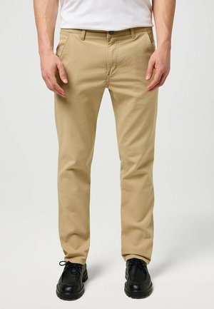 Beige Baumwollhose mit geradem Schnitt, mit Reißverschluss und zwei Fronttaschen, kombiniert mit schwarzen Lederschuhen und einem weißen T-Shirt.