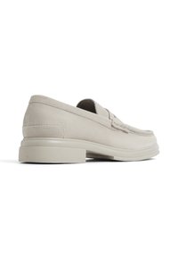 Zapatos de ante color beige claro, tipo slip-on, con un tacón bajo, que presentan costuras visibles y un diseño de punta redondeada. Textura suave y mínimos adornos.