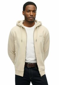 Felpa beige con zip, cappuccio regolabile, tasche frontali e logo ricamato. Abbinata a una t-shirt bianca e jeans scuri. Texture liscia.