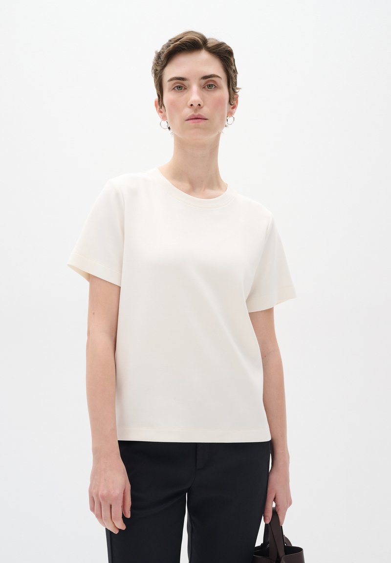 Cream short-sleeve t-shirt with a crew neckline, made of soft fabric. Worn with dark pants, model holds a small brown handbag.